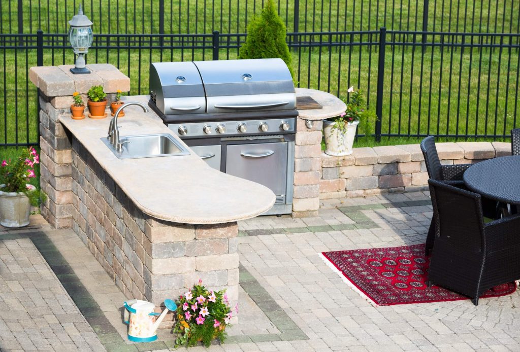 stylish outdoor kitchen on a brick patio with a built in gas barbecue,rug and dining table overlooking a green lawn and railing