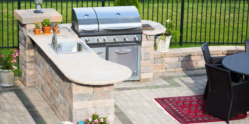 stylish outdoor kitchen on a brick patio with a built in gas barbecue,rug and dining table overlooking a green lawn and railing
